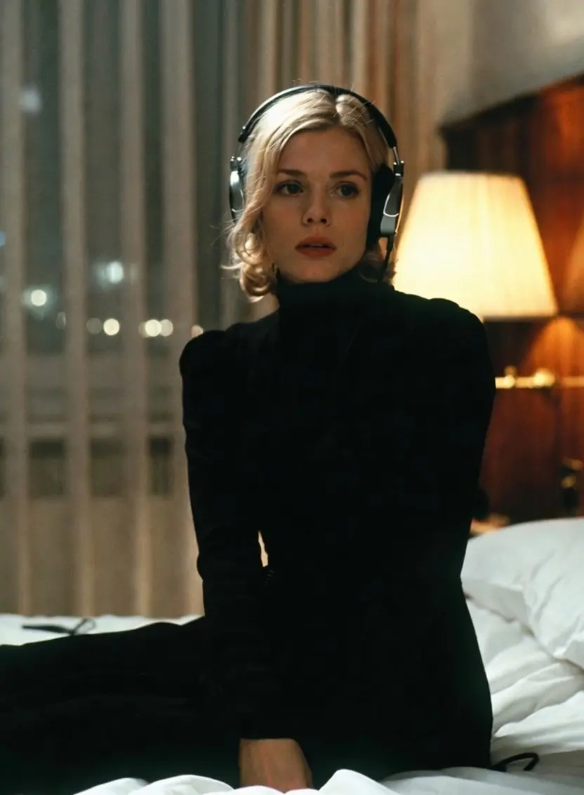 Femme blonde confiante en tenue noire élégante avec casque audio écoutant la bande originale de Atomic Blonde (2017) assise sur le bord d’un lit dans une chambre d’hôtel sombre des années 80 à Berlin.